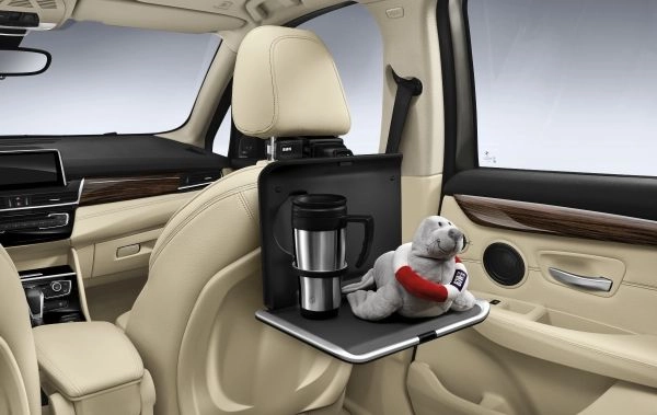 Klapptisch ist am Travel & Comfort Basisträger an der Rückseite eines Vordersitzes befestigt, darauf stehen ein Becher und ein Stofftier im Fahrzeuginnenraum