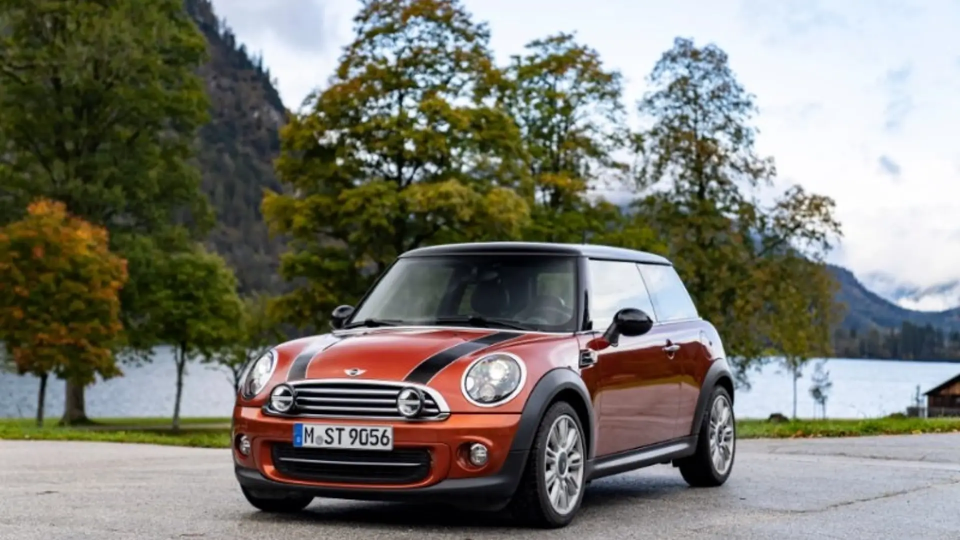 Oranger MINI Cooper der dritten Generation steht auf einem Parkplatz vor See und Bäumen