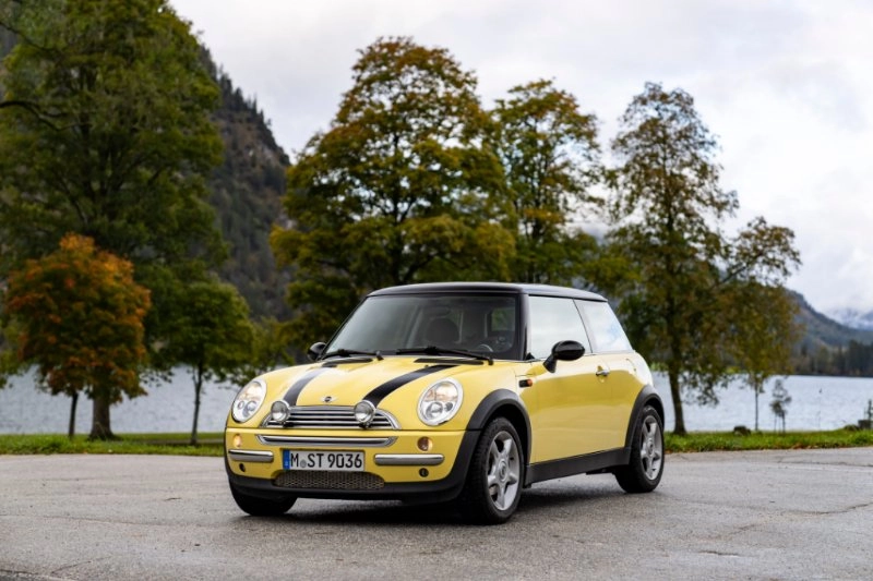 Gelber MINI Cooper der zweiten Generation steht auf einem Parkplatz vor Bäumen und See