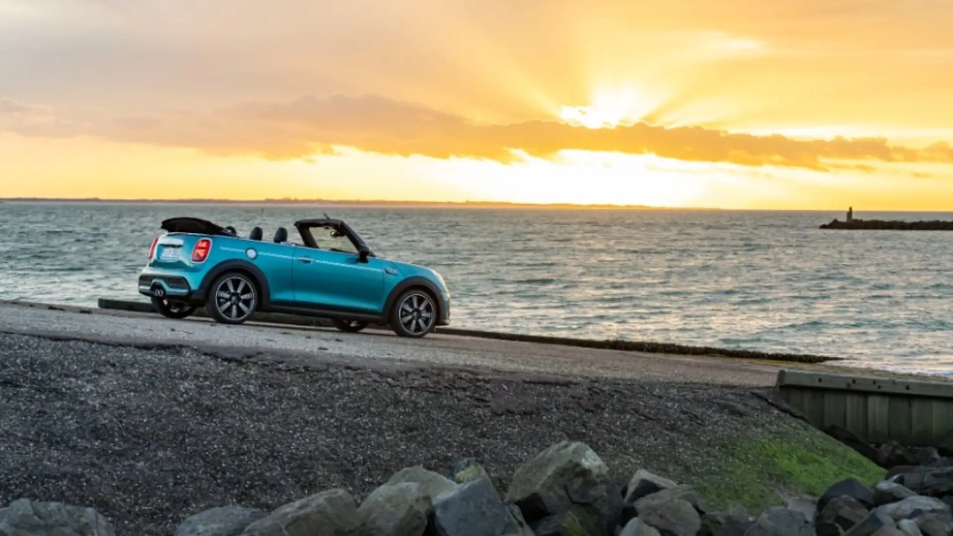 Türkiser Mini Cooper Cabrio welcher mit offenem Dach am Strand vor dem Sonnenuntergang steht