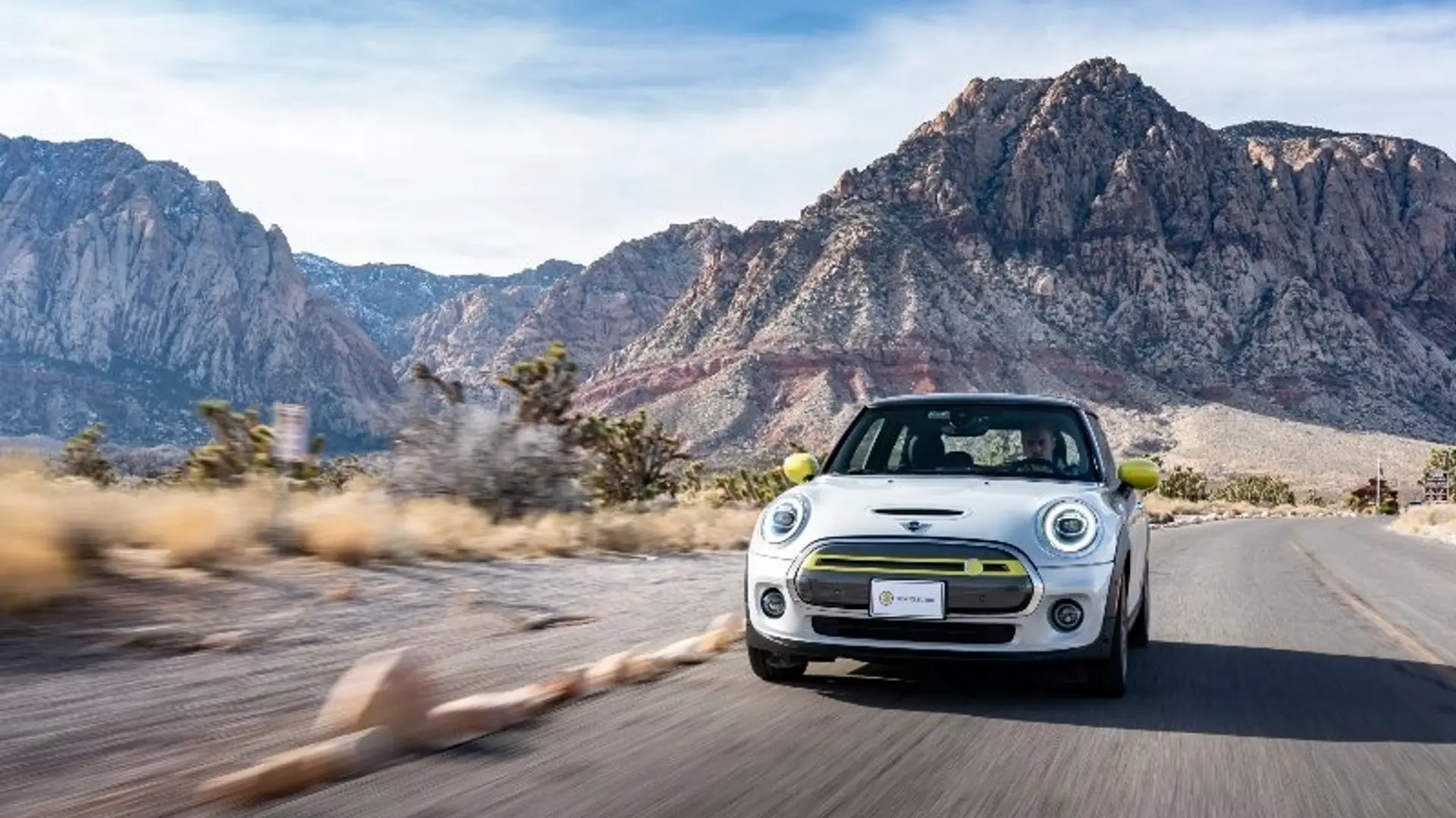 Weißer MINI Cooper fährt auf einer Landstraße durch eine Wüstenlandschaft, im Hintergrund sind Berge zu sehen