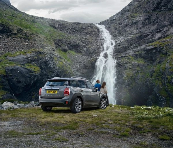Grauer MINI Countryman steht vor einem Wasserfall in einer bergigen Landschaft, zwei Personen lehnen sich gegen die Motorhaube