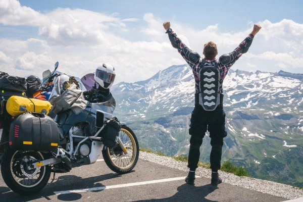Man steht vor einem Tal und hebt die Arme. Schwer beladenes Motorrad steht neben ihm