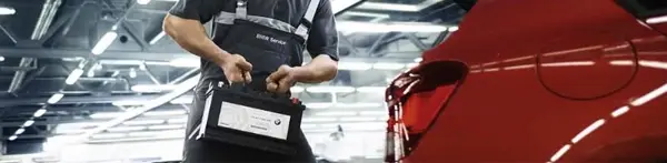 Ein Mann trägt eine Autobatterie neben einem roten BMW in einer Werkstatt