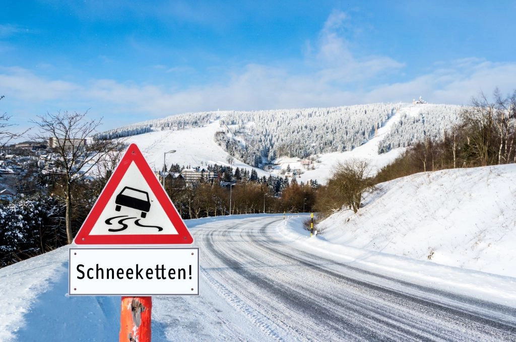 Verkehrsschild mit Hinweis auf Schneekettenpflicht steht am verschneiten Straßenrand, im Hintergrund sind eine schneebedeckte Berglandschaft und eine winterliche Straße zu sehen