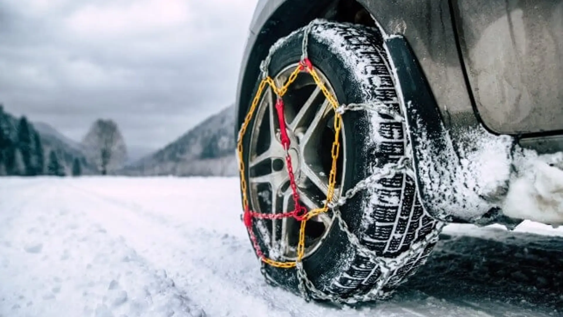 Autoreifen mit montierten Schneeketten steht auf verschneiter Straße, im Hintergrund sind winterliche Landschaft und schneebedeckte Fläche